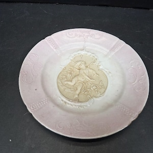 May include: A round, antique plate with a light pink rim and a white center. The plate features a raised relief design of a cherub and floral elements. The rim has a decorative pattern. The plate is on a dark surface.