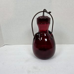 May include: A deep red glass decorative bottle with a dark metal handle. The bottle has a bulbous body and a narrower neck with a decorative stopper. The handle arches over the top, connecting to the bottle.
