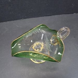 Op de afbeelding: Een heldere glazen serveerschaal met een groene rand en handvat, geaccentueerd met gouden rand en decoratieve etsen. De schaal heeft een unieke vorm en een kleine, ronde basis. Het handvat is ook van helder glas.