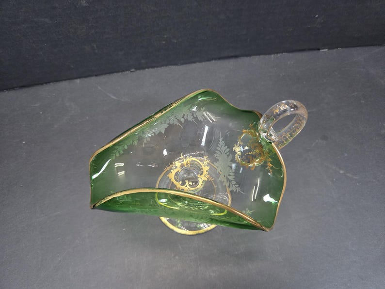 Op de afbeelding: Een heldere glazen serveerschaal met een groene rand en handvat, geaccentueerd met gouden rand en decoratieve etsen. De schaal heeft een unieke vorm en een kleine, ronde basis. Het handvat is ook van helder glas.