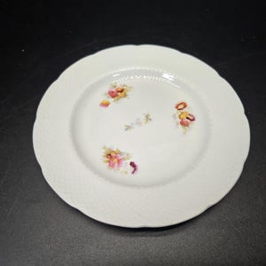 Puede incluir: Plato de cerámica blanco con un borde elevado y un diseño floral. El plato tiene un fondo blanco con un delicado patrón floral en tonos de rosa, amarillo y naranja.