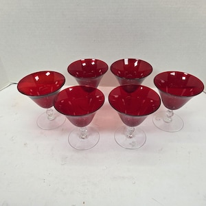 May include: Six red martini glasses with clear stems are arranged on a white surface. The glasses have a deep red bowl and a clear, delicate stem. The glasses are evenly spaced, showcasing their elegant design.