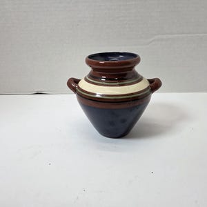 May include: A small, handcrafted ceramic vase with a dark blue base and a brown top. The vase features horizontal stripes in cream and green. It has two small handles and a dark blue interior.