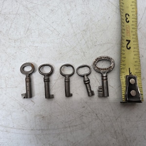 May include: Five antique metal keys of varying sizes are arranged on a light-coloured surface next to a yellow measuring tape. The keys are dark grey and have different head shapes. The measuring tape shows measurements in inches.