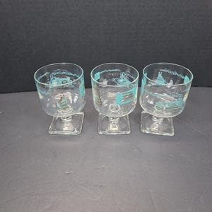 May include: Three clear glass goblets with square bases. Each goblet has a rounded bowl and is decorated with a teal-colored design around the rim. The design appears to be a vintage illustration of a building or landmark.