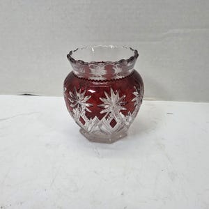 May include: A small, round glass vase featuring a deep red colour with clear, cut-glass starburst and diamond patterns. The rim has a scalloped edge, and the base is clear glass, showcasing a contrast between the vibrant red body and the transparent details.