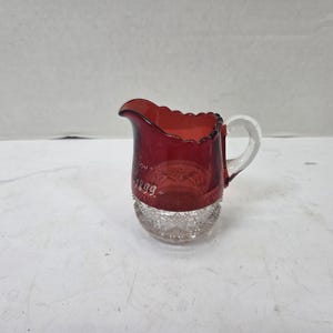 May include: A small glass creamer featuring a deep red upper section with a scalloped rim and a clear, textured base with a diamond pattern. The red glass is hand-painted with the white text 1899 and includes a clear glass handle on the side.