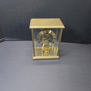 以下が含まれることがあります： ゴールドトーンの金属フレームとすべての面に透明なガラスパネルを備えた長方形の置き時計。時計の文字盤は、黒いローマ数字と黒い針が付いた円形のゴールドトーンのダイヤルで構成されており、透明なハウジング内に吊り下げられています。