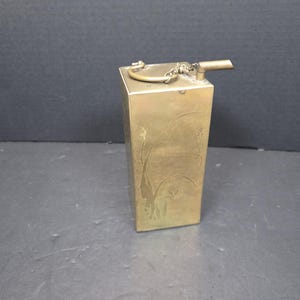 May include: A rectangular brass container with a spout and hinged lid. The container has etched floral designs and Japanese characters. The spout is connected to the lid by a small chain.