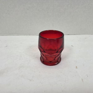 Puede incluir: Un vaso de vidrio rojo rubí con un diseño facetado. El vaso tiene forma cilíndrica con un borde ligeramente ensanchado y un patrón geométrico texturizado en la parte inferior. El vaso está vacío y se encuentra sobre una superficie blanca.