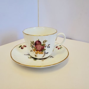 Puede incluir: Una taza de té y un platillo blancos con un borde dorado. La taza y el platillo presentan un diseño floral con frutas rojas, moradas y amarillas.