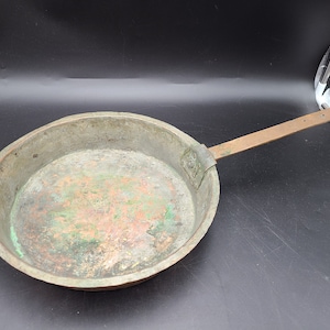 Puede incluir: Una sartén de cobre vintage con un mango largo. La sartén tiene una pátina verde y está ligeramente abollada.