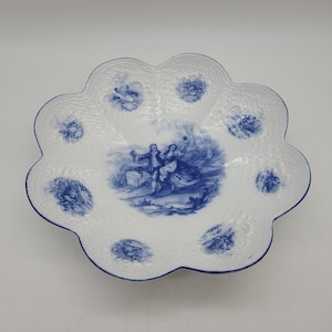 Puede incluir: Un tazón de porcelana blanca con un diseño floral azul y blanco. El tazón tiene un borde festoneado y está decorado con una escena central de una pareja en un jardín.