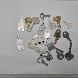 May include: A collection of various metal keys in different shapes and sizes, scattered on a white surface. The keys are in shades of silver and gold, with some showing signs of wear.