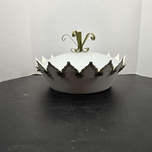 May include: White decorative bowl with a lid. The bowl has a scalloped edge with green and gold accents. The lid is topped with a green and gold handle. The bowl is sitting on a black surface.