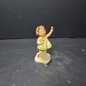 May include: A ceramic figurine of a young girl in a green dress, holding a flower and a donut. The figurine has a tag that reads "M.I. Hummel". The girl has blonde hair and is standing on a white base.