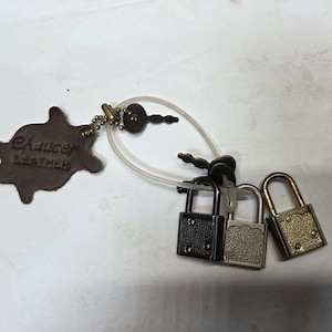 May include: A selection of miniature padlocks in black, silver, and gold, accompanied by keys. A brown leather tag, embossed with "Chaucer Leather", is attached via a clear plastic loop.