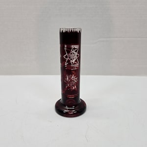 Puede incluir: Un pequeño jarrón de vidrio transparente con un tono rojo intenso. El jarrón tiene un cuerpo cilíndrico con un diseño en forma de estrella grabado en el vidrio. El jarrón tiene una base acampanada.