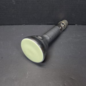 Könnte beinhalten: Eine schwarz-silberne Taschenlampe mit einer hellgrünen Linse. Der Griff ist mit einem strukturierten schwarzen Material umwickelt. Die Taschenlampe liegt auf einer dunklen Oberfläche.