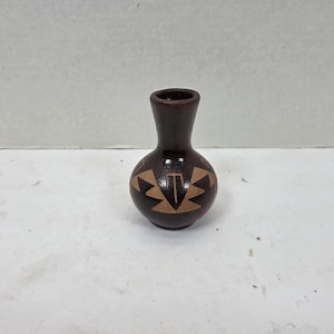 Puede incluir: Un pequeño jarrón de cerámica marrón oscuro con cuerpo redondeado y cuello estrecho. El jarrón presenta un patrón geométrico beige alrededor del cuerpo. El jarrón mide unos 10 cm de alto y es probablemente un objeto decorativo.