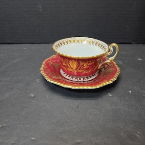 Peut inclure: Un service à thé rouge avec des détails dorés. La tasse et la soucoupe ont un bord festonné et un bord doré. La tasse présente une anse dorée et un motif floral doré. La soucoupe a un motif floral doré assorti.