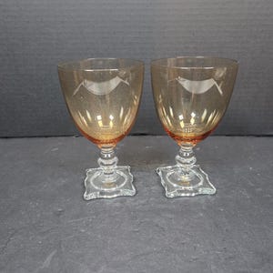 May include: Two amber-colored wine glasses with a bird design etched into the bowl. The glasses have a clear, square base and a decorative stem. The glasses are likely vintage and suitable for serving wine.