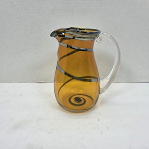 Può includere: Brocca in vetro color ambra con manico trasparente. La brocca presenta un design decorativo di vortici argentati e neri. Il bordo è accentuato da una fascia scura. Un pezzo decorativo per servire bevande.