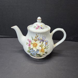 May include: A white porcelain teapot with a ribbed body and a matching lid. The teapot features a colourful floral pattern of yellow daffodils, purple irises, and small blue flowers on the front, with a smaller floral motif on the lid.