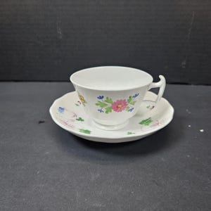 May include: White teacup and saucer set with floral design. The teacup has a curved handle and sits on a matching saucer. The floral pattern includes pink, blue, and green flowers. A classic tea set for serving tea.