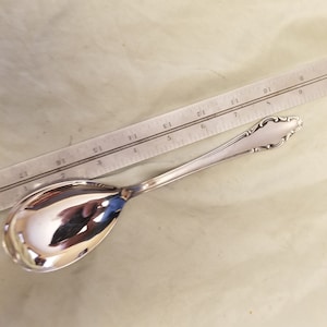 Può includere: Un cucchiaio da portata in argento con un manico decorato. Il cucchiaio è lungo circa 15 cm e ha una ciotola arrotondata.