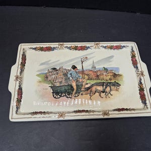Puede incluir: Bandeja rectangular de cerámica con una escena pintada a mano de una persona con un carro tirado por perros. La bandeja tiene un diseño de borde floral y de lazo. La escena representa un pueblo.