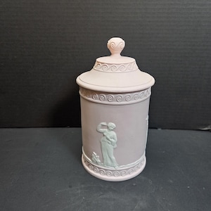 May include: A light pink ceramic jar with a lid, featuring a classical relief design. The jar has a cylindrical shape with a decorative band of swirls and a small finial on the lid. The relief depicts figures in a classical style.