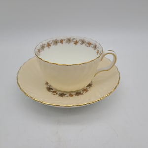 May include: Cream-colored teacup and saucer set with gold trim. The teacup has a delicate floral pattern around the rim and on the saucer. The set is made of fine china and is ideal for serving tea.