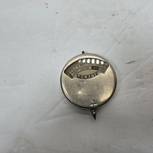 Puede incluir: Un comprobador de batería Buckeye de 6 voltios vintage. El dispositivo circular es de color plateado con una aguja y un indicador que indica "Start Charging" y "OK Stop". La palabra "Buckeye" está impresa en la cara.