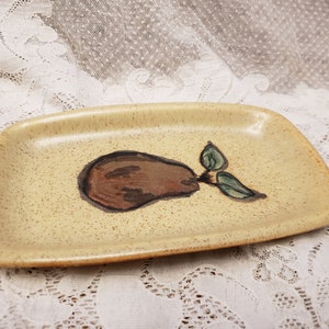 Può includere: Un piccolo piatto rettangolare in ceramica con una glassa beige macchiata. Una pera marrone dipinta a mano con foglie verdi è centrata sul piatto.