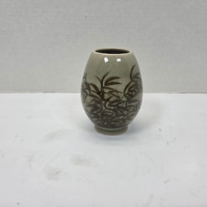 May include: A small, egg-shaped ceramic vase with a light green glaze. The vase is decorated with a dark brown bamboo leaf design. The vase has a small, round base and a narrow opening at the top. A decorative item.
