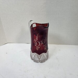 May include: A deep red glass pitcher featuring a clear glass handle and a clear, textured base with a diamond-cut pattern. The body of the pitcher displays the etched cursive text "Exile" followed by the number "1904" in a decorative script style.