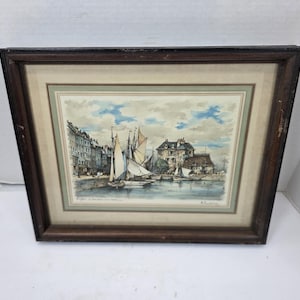 May include: Framed watercolor print of a harbor scene with sailboats, buildings, and a cloudy sky. The artwork is in a brown wooden frame with a cream-colored matting. The print depicts a coastal town.