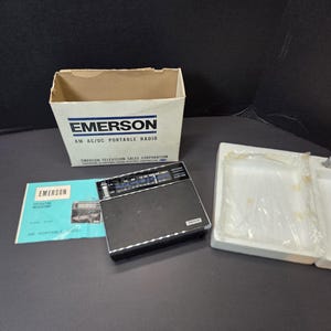 Puede incluir: Una radio portátil Emerson AM AC/DC negra con un dial plateado, acompañada de su caja de cartón blanca original etiquetada como Emerson Television Sales Corporation, un manual de instrucciones azul y blanco e inserciones de embalaje de poliestireno.