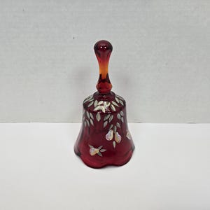 May include: A red glass bell with a white painted floral design. The bell has a handle and a scalloped edge.