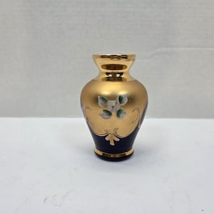 Könnte beinhalten: Eine kleine, dekorative Vase mit goldfarbener Außenseite und dunkelblauem Boden. Die Vase hat ein Blumendesign mit weißen und grünen Akzenten. Ober- und Unterseite haben einen goldenen Rand. Die Vase ist etwa 10 cm hoch.