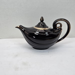 Puede incluir: Tetera de cerámica negra con detalles dorados. La tetera tiene un asa y un pico curvos, y una tapa con un pequeño adorno. El cuerpo de la tetera tiene un diseño acanalado. Un artículo decorativo para la cocina.