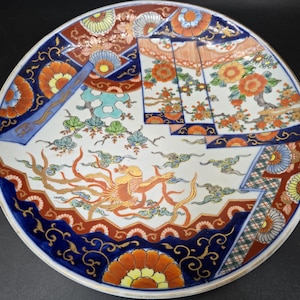 May include: A large, round, white ceramic plate with a colorful Japanese-style design. The plate features a phoenix bird, flowers, and geometric patterns in shades of blue, red, orange, and gold.