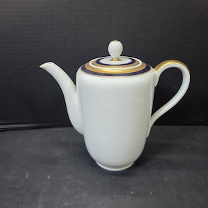 May include: A white porcelain teapot featuring a curved spout, a C-shaped handle, and a lid topped with a textured white knob. The lid is decorated with concentric bands of deep navy blue and metallic gold, providing a refined contrast to the body.