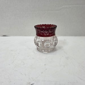 May include: A small clear glass toothpick holder with a scalloped rim and a deep red band around the top. The red section features the white painted text World's Fair 1893. The base of the glass has a repeating pattern of circular indentations.