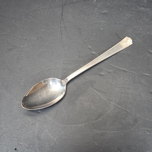 Puede incluir: Una cuchara plateada con un cuenco redondeado y un mango rectangular. La cuchara está hecha de un metal brillante y reflectante y está colocada sobre una superficie gris oscuro. Es un utensilio común para comer.