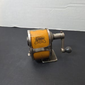 May include: Vintage yellow and silver pencil sharpener with the word "Giant" printed on the side. The sharpener has a hand crank and a small base. The text "Automatic Pencil Sharpener" is also visible.