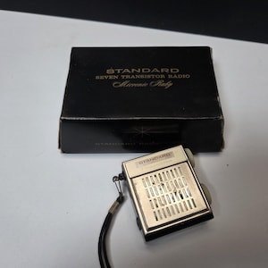 May include: A vintage silver-toned transistor radio with a black wrist strap and a black box. The box is labeled "STANDARD SEVEN TRANSISTOR RADIO Micronic Ruby". The radio has a speaker grill and two gray dials.