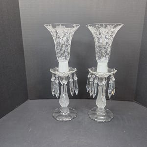 Puede incluir: Dos candelabros de cristal transparente con diseños florales grabados. Cada soporte tiene una parte superior acampanada, una base decorativa y detalles de cristal colgantes. Los candelabros se exhiben sobre una superficie oscura.