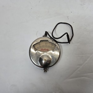 May include: A vintage Sterling ammeter with a silver-colored, circular casing. The gauge displays numbers from 0 to 30, with the word "Amperes" above and "Sterling" below. A black wire is attached to the top.